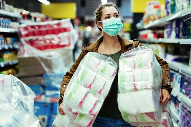 Kostenloses Foto frau, die eine schützende gesichtsmaske trägt, während sie toilettenpapierpakete trägt und in zeiten einer viruspandemie kauft