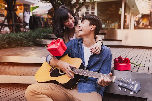 Frau, die einen Mann umarmt und halten ein Geschenk, während er die Gitarre spielt