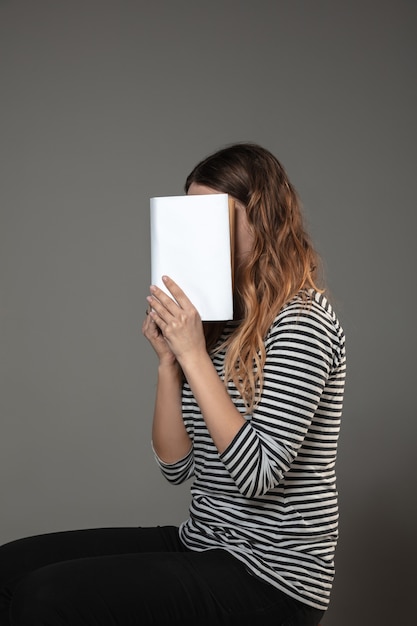 Frau, die gesicht mit buch bedeckt, während sie an grauer wand liest. feiern, bildung, kunst, neues charakterkonzept genießen.