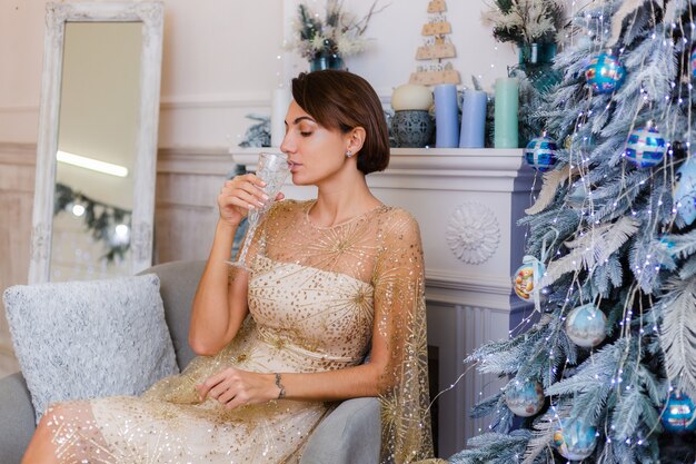 Frau, die glänzendes goldenes Abendweihnachtskleid hält, das Glas Champagner durch blauen Jahrbaum zu Hause hält