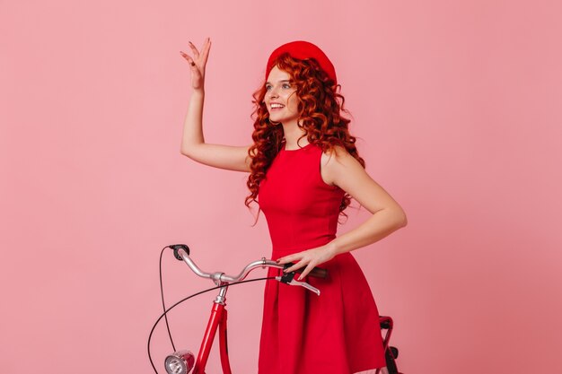 Frau, die Gruß beim Sitzen auf dem Fahrrad auf rosa Raum winkt. Dame in roter Baskenmütze und Kleid.