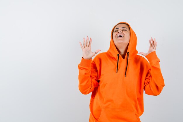 Frau, die Hände hebt, mit geschlossenen Augen in orangefarbenem Hoodie aufschaut und glücklich aussieht