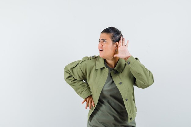 Frau, die Hand nahe Ohr in Jacke, T-Shirt hält und neugierig schaut.