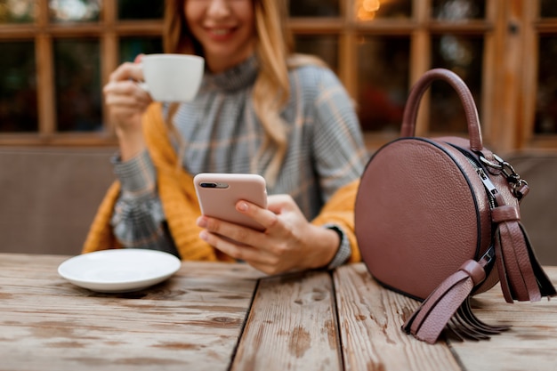 Frau, die Handy, SMS und Kaffeetrinken verwendet. Stilvolle Tasche auf dem Tisch. Trägt graues Kleid und orange Plaid. Genießen Sie einen gemütlichen Morgen im Café.