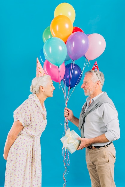 Frau, die ihren Ehemann hält bunte Ballone und Geburtstagsgeschenk auf blauem Hintergrund betrachtet