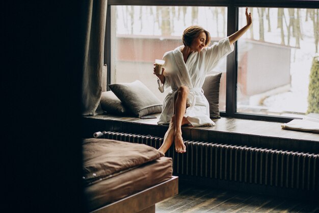 Frau, die Kaffee im Bademantel durch das Fenster zu Hause trinkt