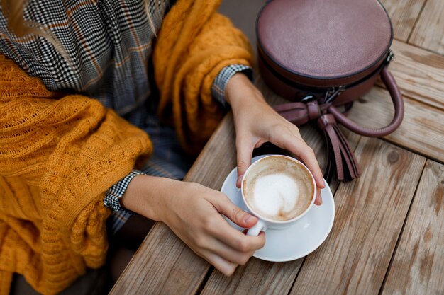 Frau, die Kaffee trinkt. Stilvolle Tasche auf dem Tisch. Trägt graues Kleid und orange Plaid. Genießen Sie einen gemütlichen Morgen im Café.