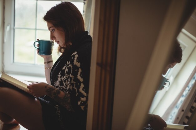 Frau, die Kaffee trinkt und ein Buch liest, während sie am Fensterbrett sitzt