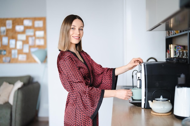 Frau, die Kaffeemaschine verwendet, um große Tasse Kaffee zu Hause zu machen Frau trägt Seidenrobe zu Hause, während sie einen Latte zubereitet