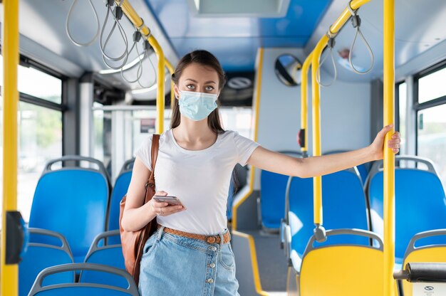 Frau, die mit dem öffentlichen Bus mit dem Smartphone reist, während sie eine medizinische Maske zum Schutz trägt