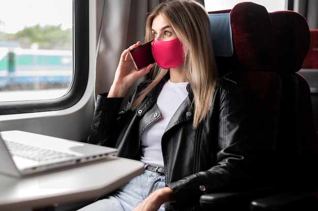 Frau, die mit dem Zug reist und telefoniert, während sie eine medizinische Maske trägt und am Laptop arbeitet