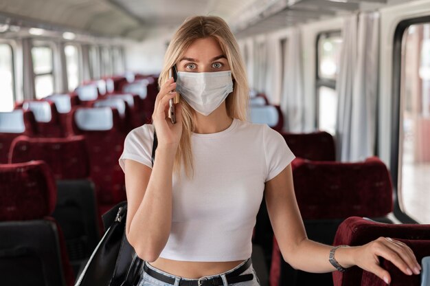 Frau, die mit dem Zug reist und telefoniert, während sie eine medizinische Maske trägt