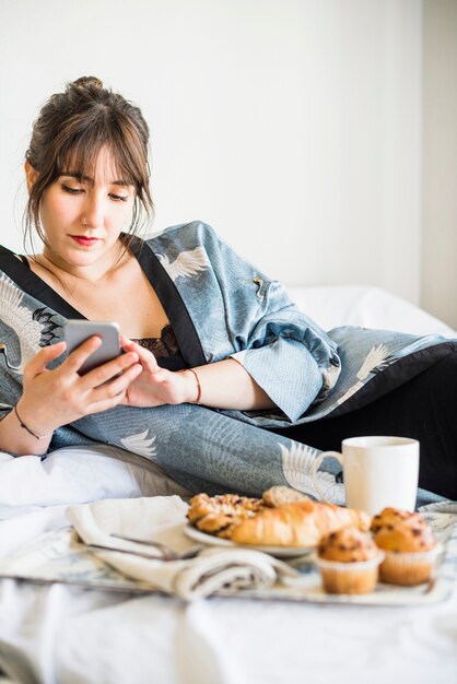 Frau, die Mobiltelefon mit Frühstück auf Bett verwendet
