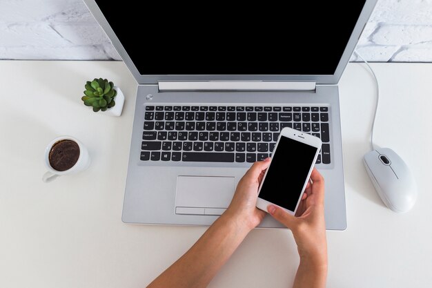 Frau, die Mobiltelefon über dem Laptop auf dem Schreibtisch verwendet