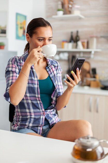 Frau, die morgens aromatischen Tee trinkt, der auf dem Smartphone surft