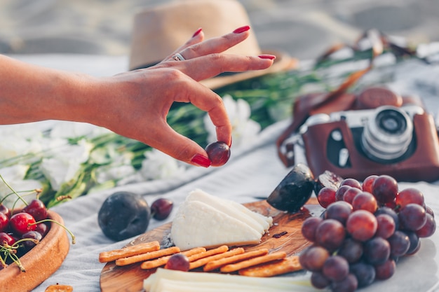 Frau, die Obst mit Käse und Früchten auf hölzernem Schneidebrett und Kamera, Hut und Blumen im Strand hält.