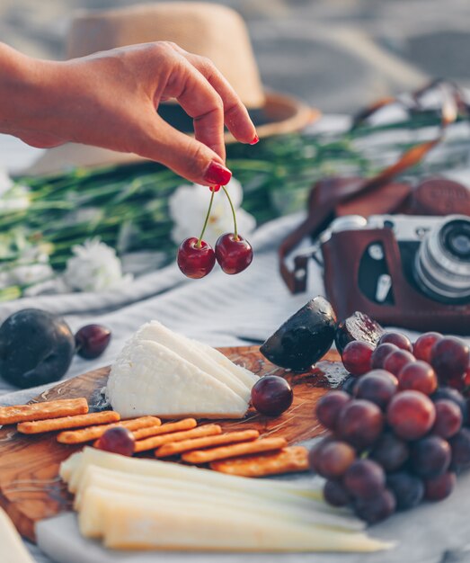 Frau, die Obst mit Käse und Früchten auf hölzernem Schneidebrett und Kamera, Hut und Blumen im Strand hält.