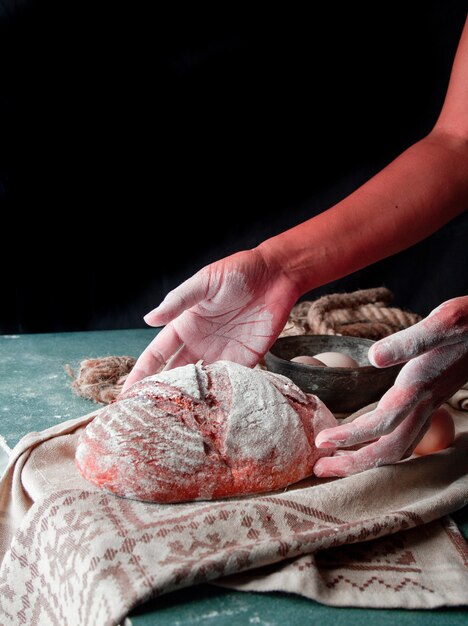 Frau, die selbst gemachtes rundes Brot mit Mehl auf die Hände und auf die Oberseite des Brotes auf ein rustikales Tuch setzt.