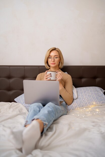 Frau, die sich entspannt und Tasse heißen Kaffee oder Tee mit Laptop-Computer im Schlafzimmer trinkt.