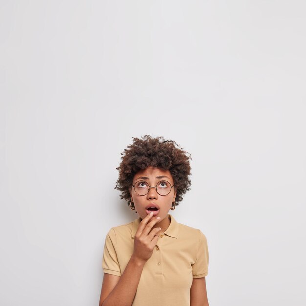 Frau, die sich oben konzentriert, hält den Mund geöffnet, fühlt sich erschreckt, kann nicht an etwas Schockierendes glauben, trägt transparente Brille braunes lässiges T-Shirt isoliert auf weiß. Oh nein, wie beängstigend