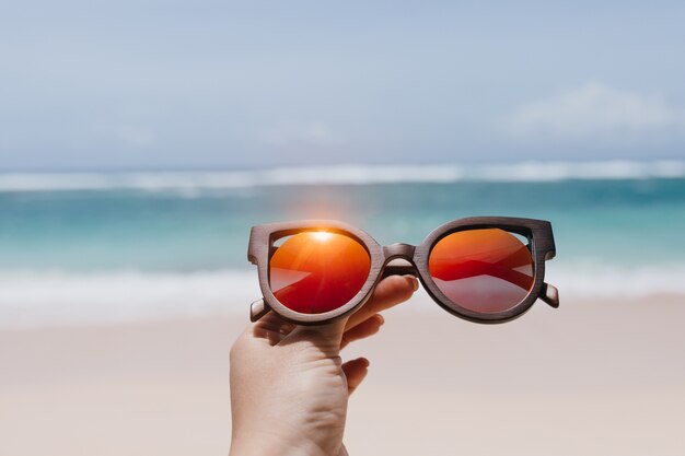 Frau, die stilvolle Sommersonnenbrille über Meer hält. Außenfoto der weiblichen Hand mit den Gläsern am Strand.