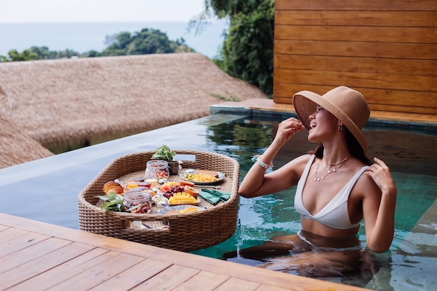 Frau, die tropisches gesundes Frühstück an der Villa auf schwimmendem Tisch hat