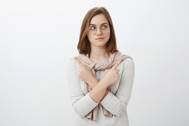 Frau, die versucht, wählte welches Produkt besser. Unsichere entspannte und kluge kreative Frau mit kurzen braunen Haaren in der Brille, die richtig schaut, während sie die Arme auf der Brust verschränkt, die auf beiden Seiten unsicher zeigen