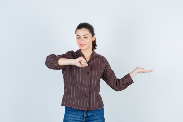 Frau, die vorgibt, auf die Uhr am Handgelenk zu schauen, die Handfläche in einem braun gestreiften Hemd zur Seite spreizt und nachdenklich aussieht, Vorderansicht.