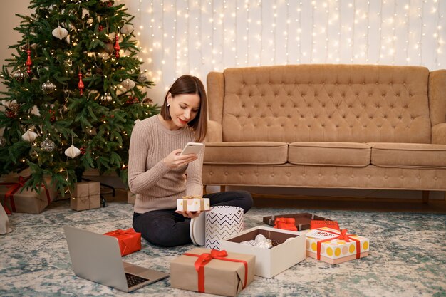 Frau, die weihnachtsgeschenk einwickelt und bilder am telefon beim sitzen in weihnachten macht