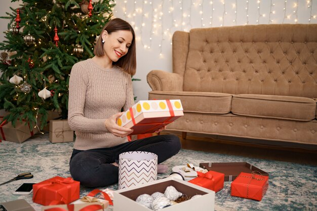 Frau, die Weihnachtsgeschenk verpackt, während sie auf dem Teppich im Wohnzimmer sitzt