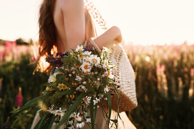 Frau, die Wildflowersblumenstrauß im Strohbeutel, gehend in Blumenfeld auf Sonnenuntergang hält.