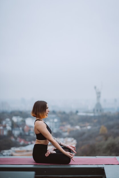 Frau, die Yoga auf der Matte auf dem Dach praktiziert und Yogaübungen macht