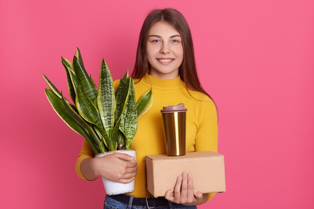 Frau, die zu neuem Haus mit Kartonschachtel, Kaffee oder Tee zum Mitnehmen und weißem Blumentopf mit Sansevieria zieht