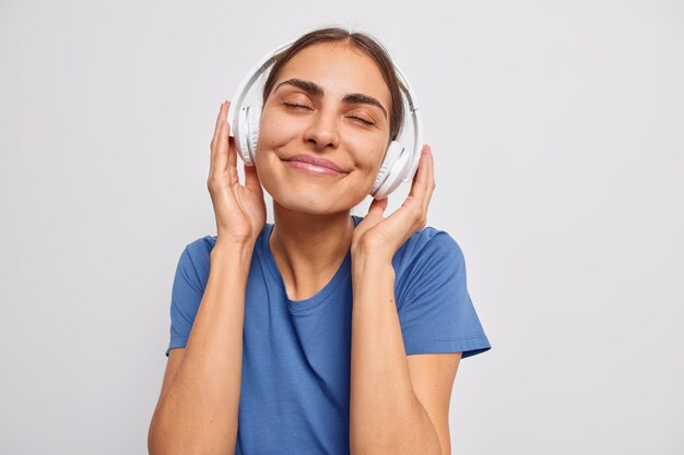 Frau genießt angenehme Melodie in kabellosen Kopfhörern fängt jedes Liedchen ein hält die Augen geschlossen in einem lässigen blauen T-Shirt auf Weiß gekleidet