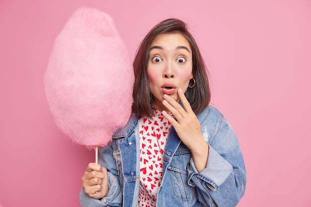 Kostenloses Foto frau genießt es, lieblingssüße zu essen, hält appetitanregende zuckerwatte auf dem stock, die fassungslos ist, um herauszufinden, wie kalorien verbraucht werden, hat im park in jeansjacke gekleidet. leckeres dessert