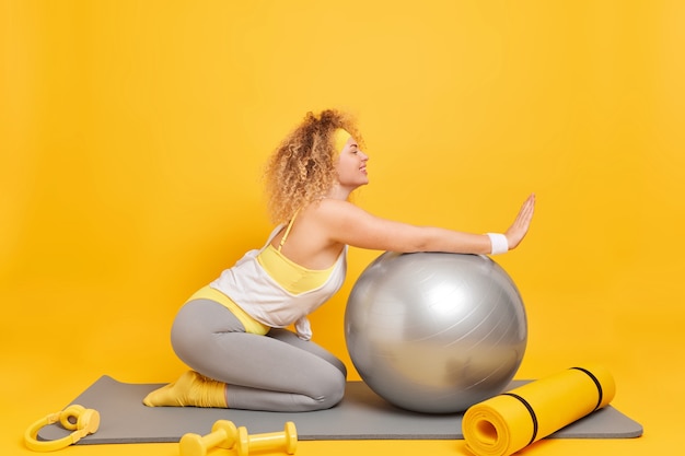 Frau genießt fitnesstraining zu hause lehnt sich an schweizer ballposen auf der matte mit karemat-hanteln und kopfhörern isoliert auf gelb