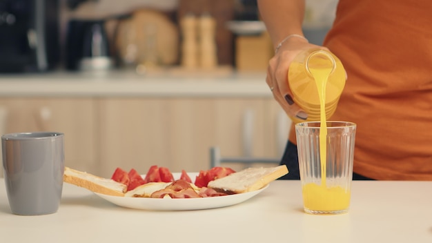 Frau gießt frischen Saft in Glas zum Frühstück in der Küche. Frau, die gesunden und natürlichen Orangensaft trinkt. Hausfrau trinkt gesunden, natürlichen, hausgemachten Orangensaft. Erfrischender Sonntagmorgen