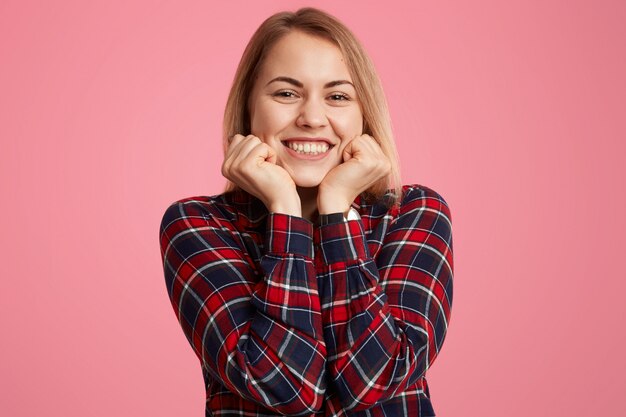 Frau hält Kinn, lächelt breit, zeigt weiße perfekte Zähne, ist gut gelaunt, lässig gekleidet