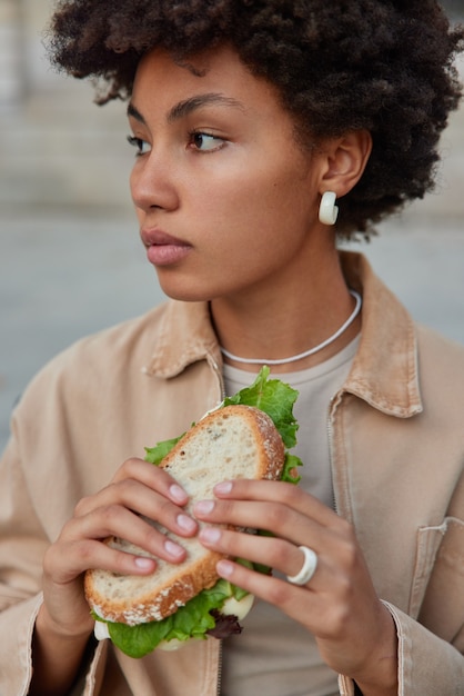 Frau hat Snack auf der Straße hält leckeres Sandwich isst leckeres Fast Food sieht beiseite nachdenklich gekleidet in modischer Kleidung posiert draußen