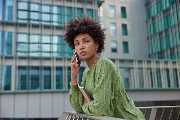 Frau hat telefongespräche über wochenendpläne während des roaming-mobilfunks trägt lässige grüne pullover-posen gegen moderne stadtgebäude