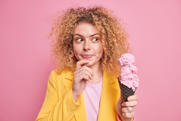 Frau hat verträumten Ausdruck hält die Hand am Kinn sieht appetitliches Eis an, das leckeres Sommerdessert isst, trägt eine formelle gelbe Jacke, die auf rosa Wand isoliert ist