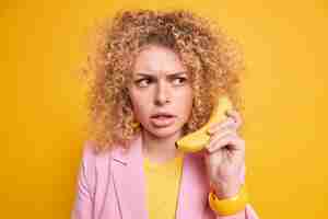 Kostenloses Foto frau hat wütenden strengen ausdruck spielt die rolle des chefs hält banane in der nähe des ohrs und gibt vor, ein telefonat zu haben, das sich in einer stilvollen jacke konzentriert