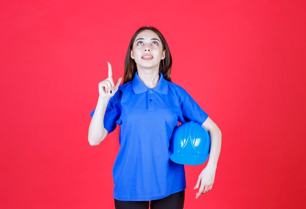 Frau im blauen Hemd, die einen blauen Helm hält und auf irgendwo herum zeigt.