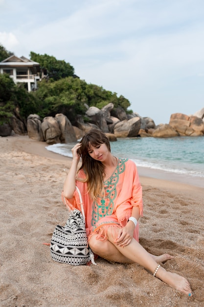 Frau im Boho-Sommerkleid, das auf Sand nahe Meer sitzt. Tropische Stimmung.