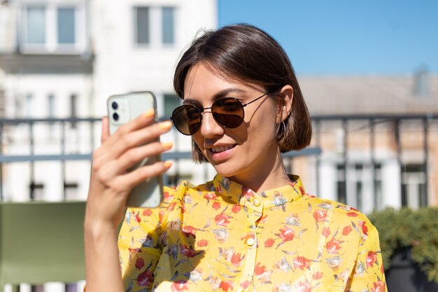 Frau im gelben Kleid auf Terrasse im Sommercafé mit Handy am sonnigen Tag