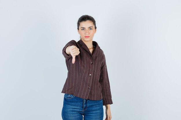 Frau im hemd, jeans, die daumen nach unten zeigt und unzufrieden aussieht, vorderansicht.