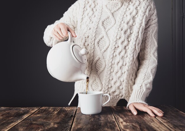 Frau im kuscheligen warmen weißen dicken Strickpullover gießt heißen Tee von der großen Keramik-Teekanne zur leeren Tasse
