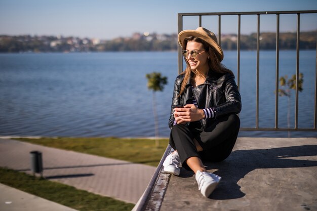 Frau im Lederjacken-Sitz auf dem Pier nahe dem See im kalten sonnigen Herbsttag