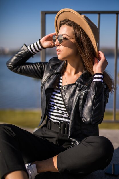 Frau im Lederjacken-Sitz auf dem Pier nahe dem See im kalten sonnigen Herbsttag