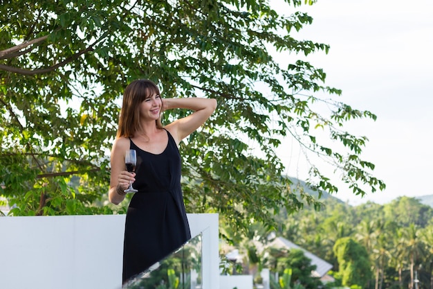 Frau im schwarzen Abendkleid mit Glas Wein auf tropischem Balkon
